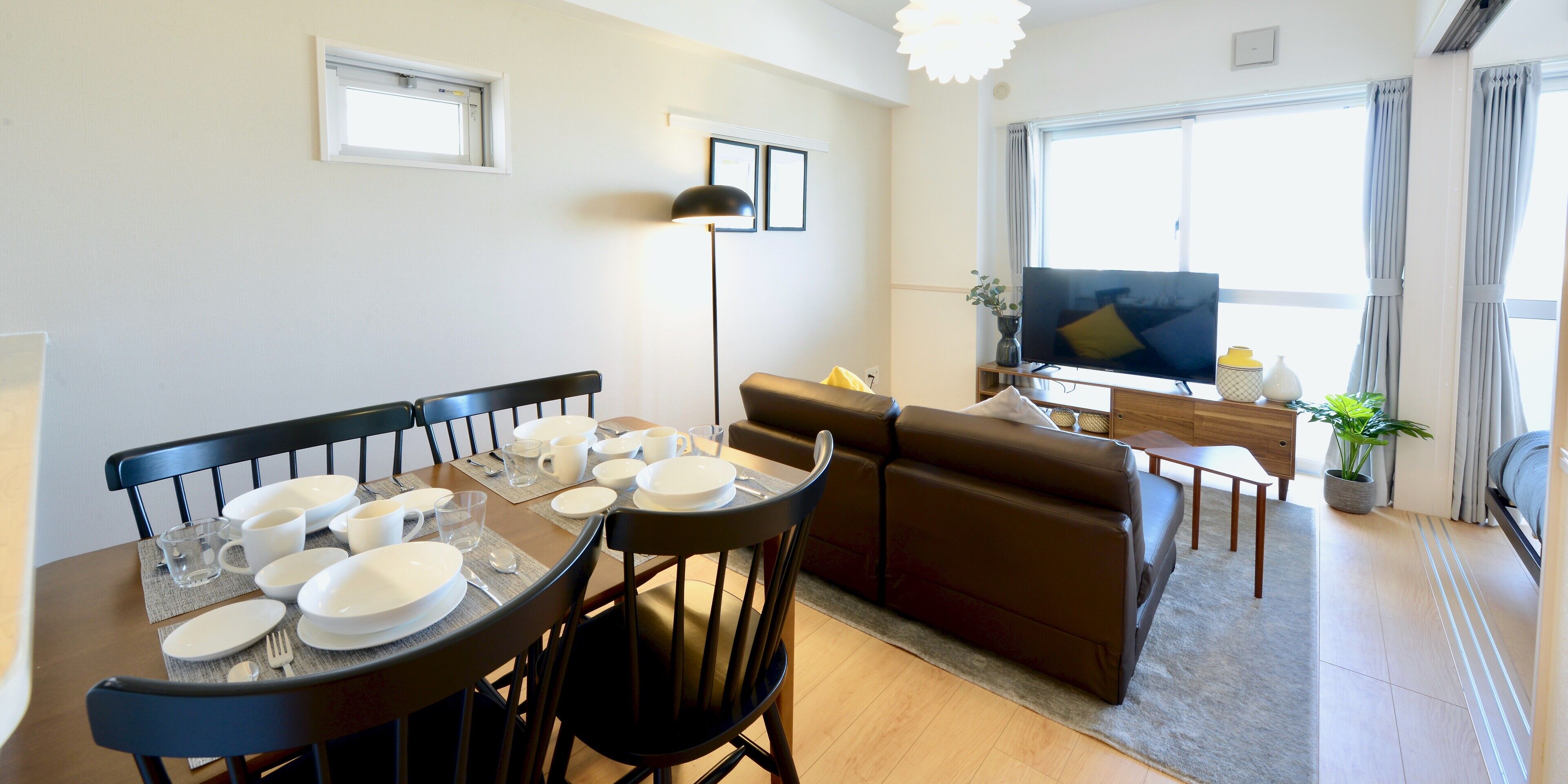 A dining area with a round table set for four, a brown leather sofa, a TV, and a window with curtains.
