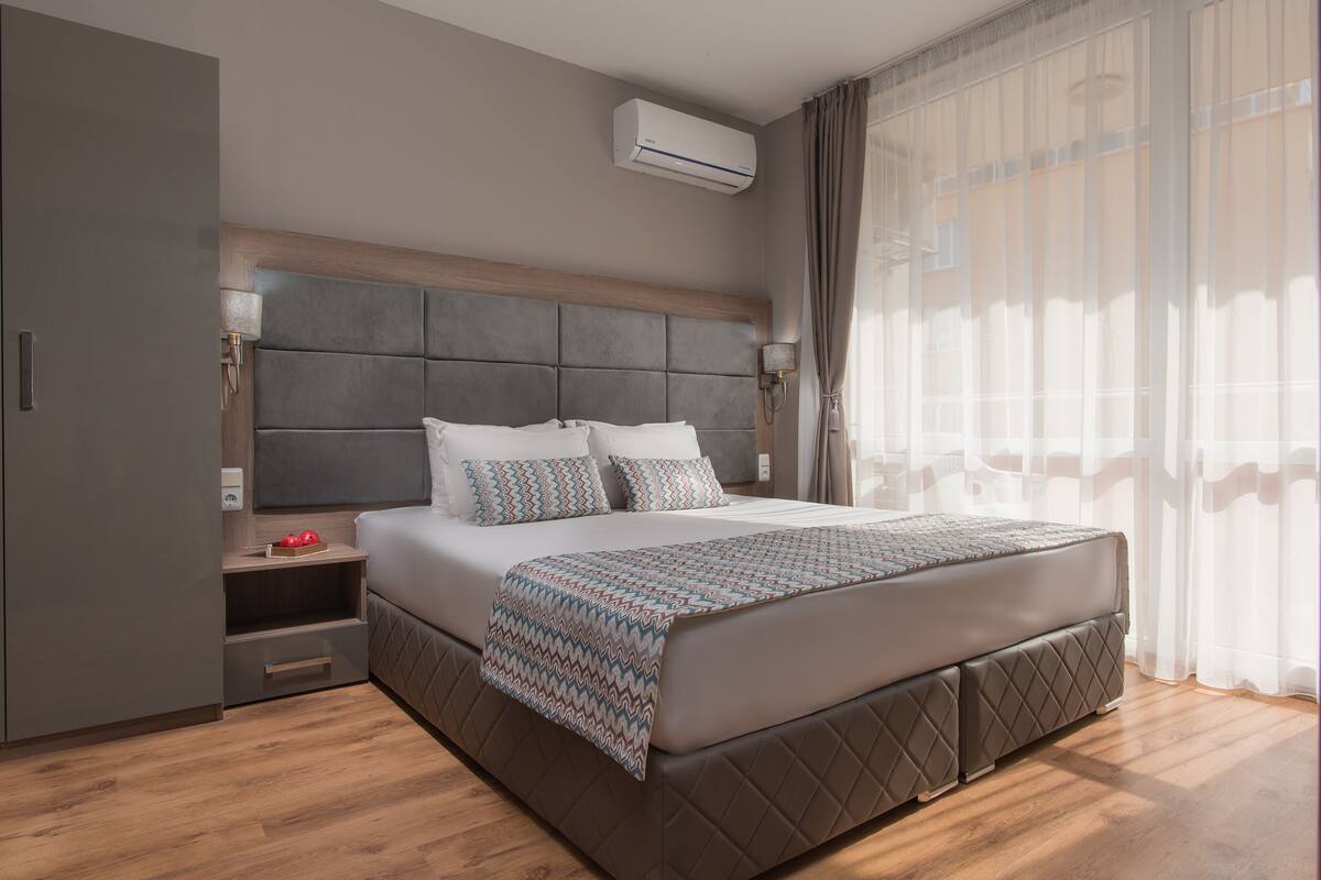 A modern hotel room with a large bed, a wardrobe, a bedside table, and an air conditioning unit.