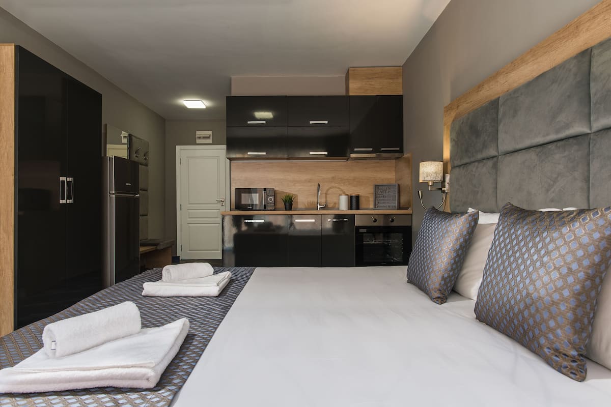 A compact hotel room with a kitchenette, a bed with patterned pillows, and a large window.