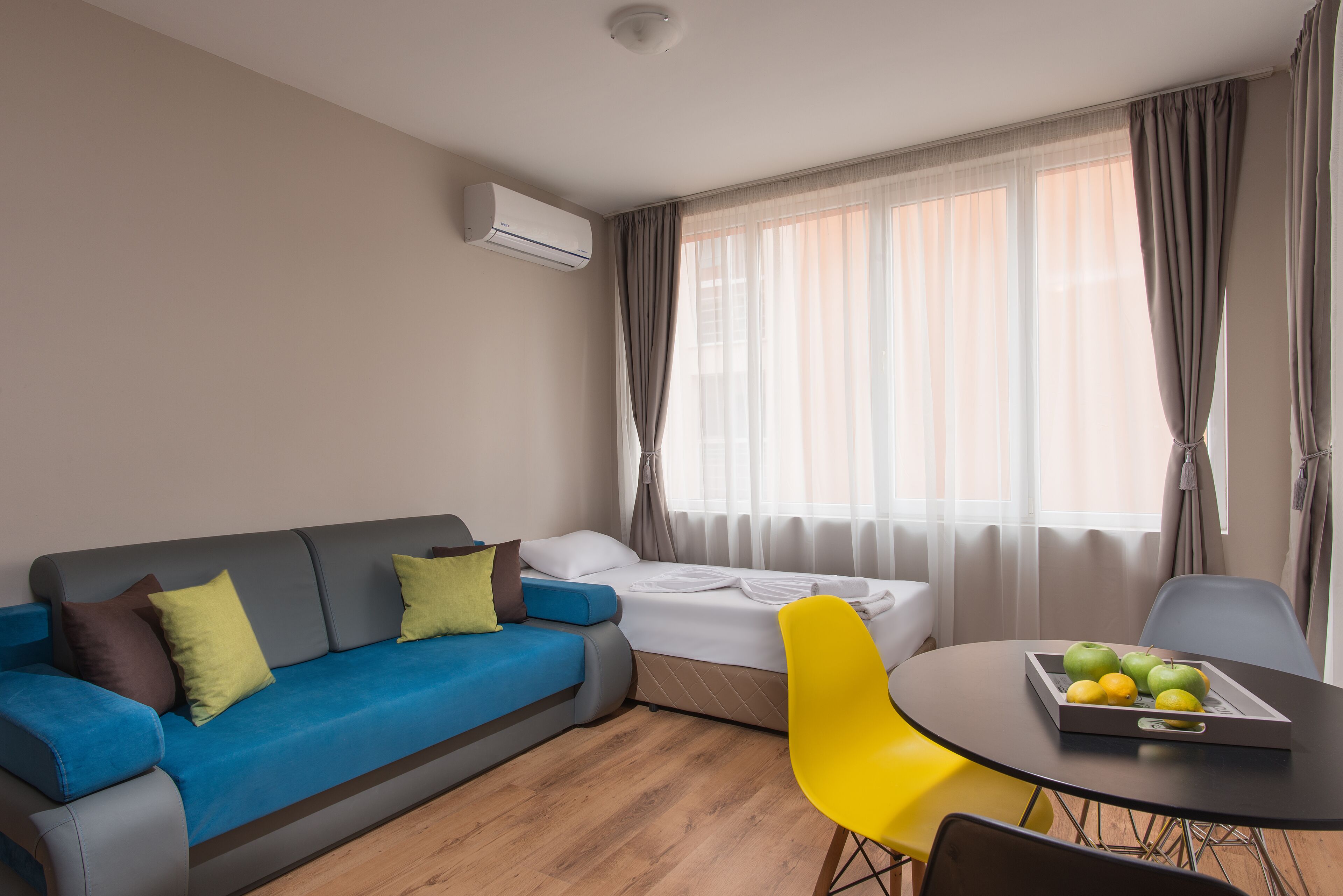 A modern hotel room with a sofa, a bed, a dining table with fruit, and a window with curtains.