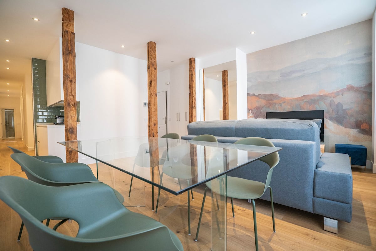 A modern living room with a glass table, green chairs, a grey sofa, and a large abstract painting on the wall.