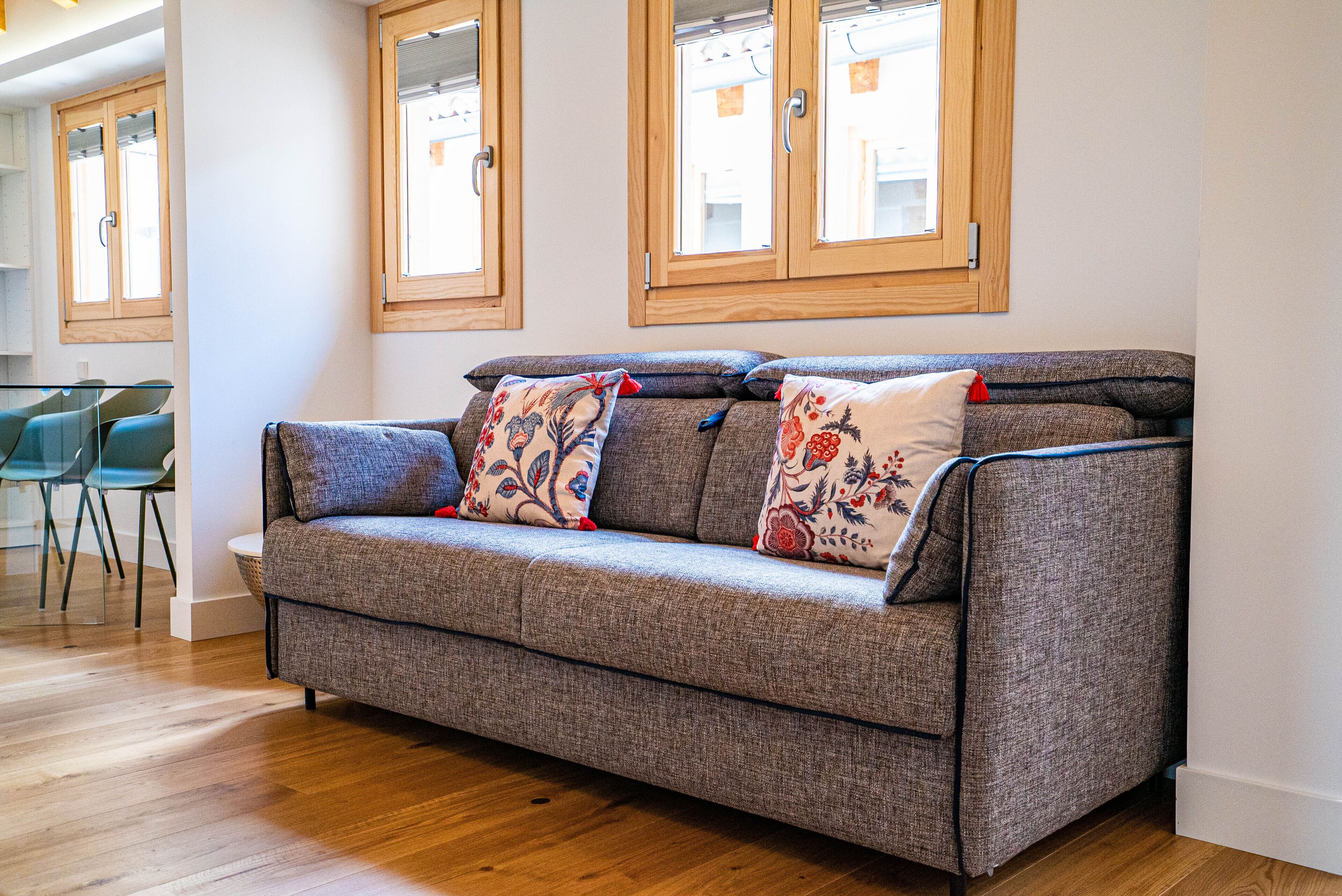 A grey sofa with floral cushions, wooden floor, and a window with wooden frames.
