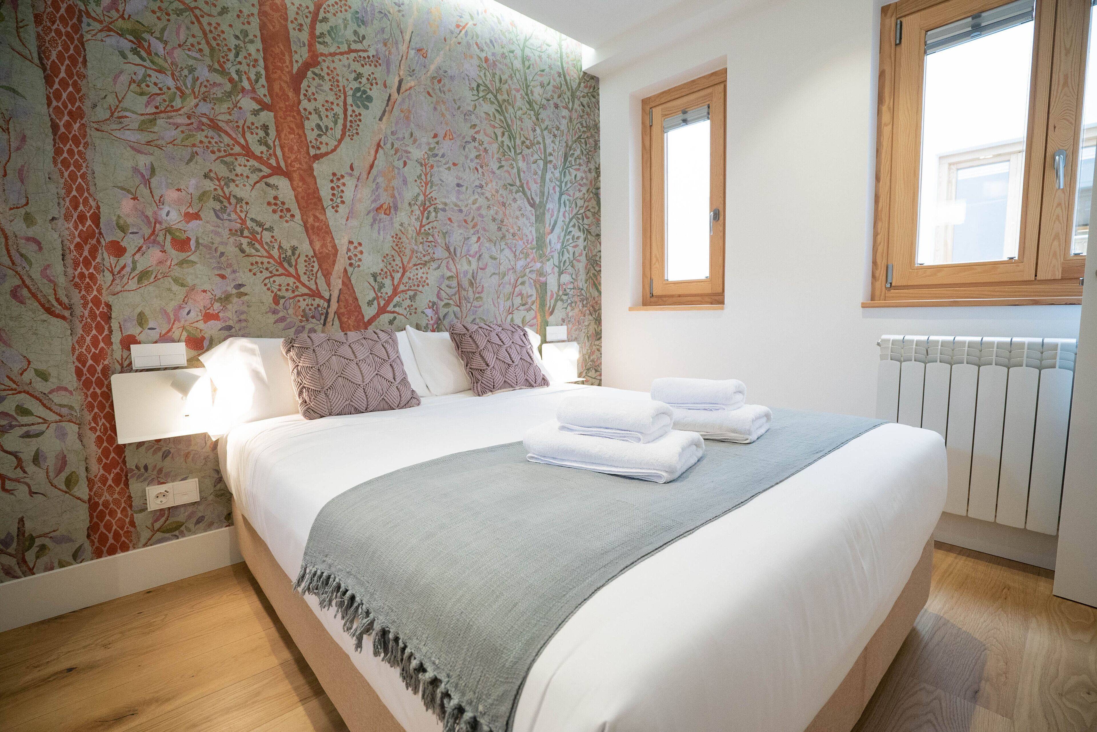 Una cama bien hecha con sábanas blancas y un plaid gris, cabecera de madera y diseño de papel tapiz floral.