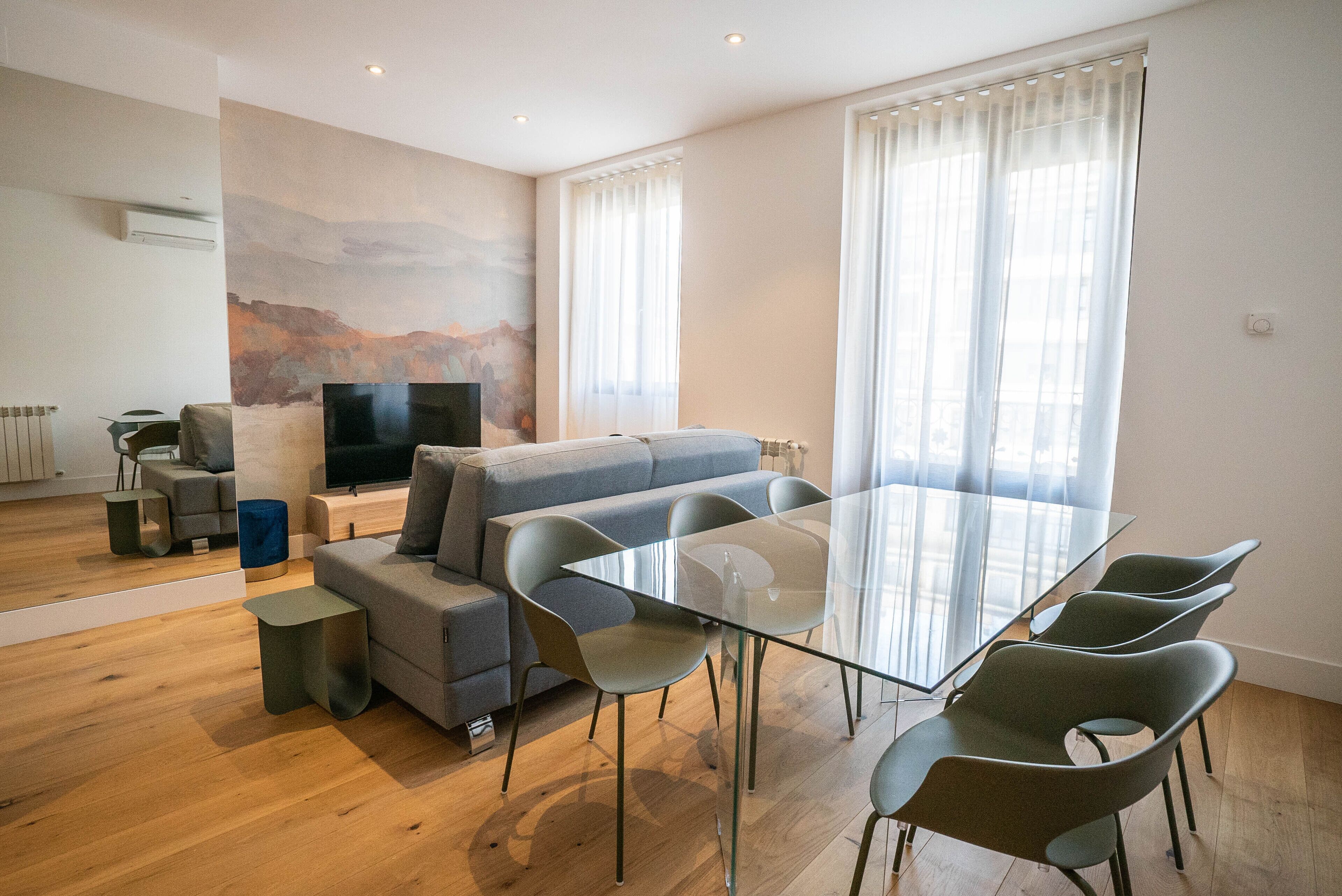 A modern living room with a glass dining table, grey sofa, and a flat-screen TV.