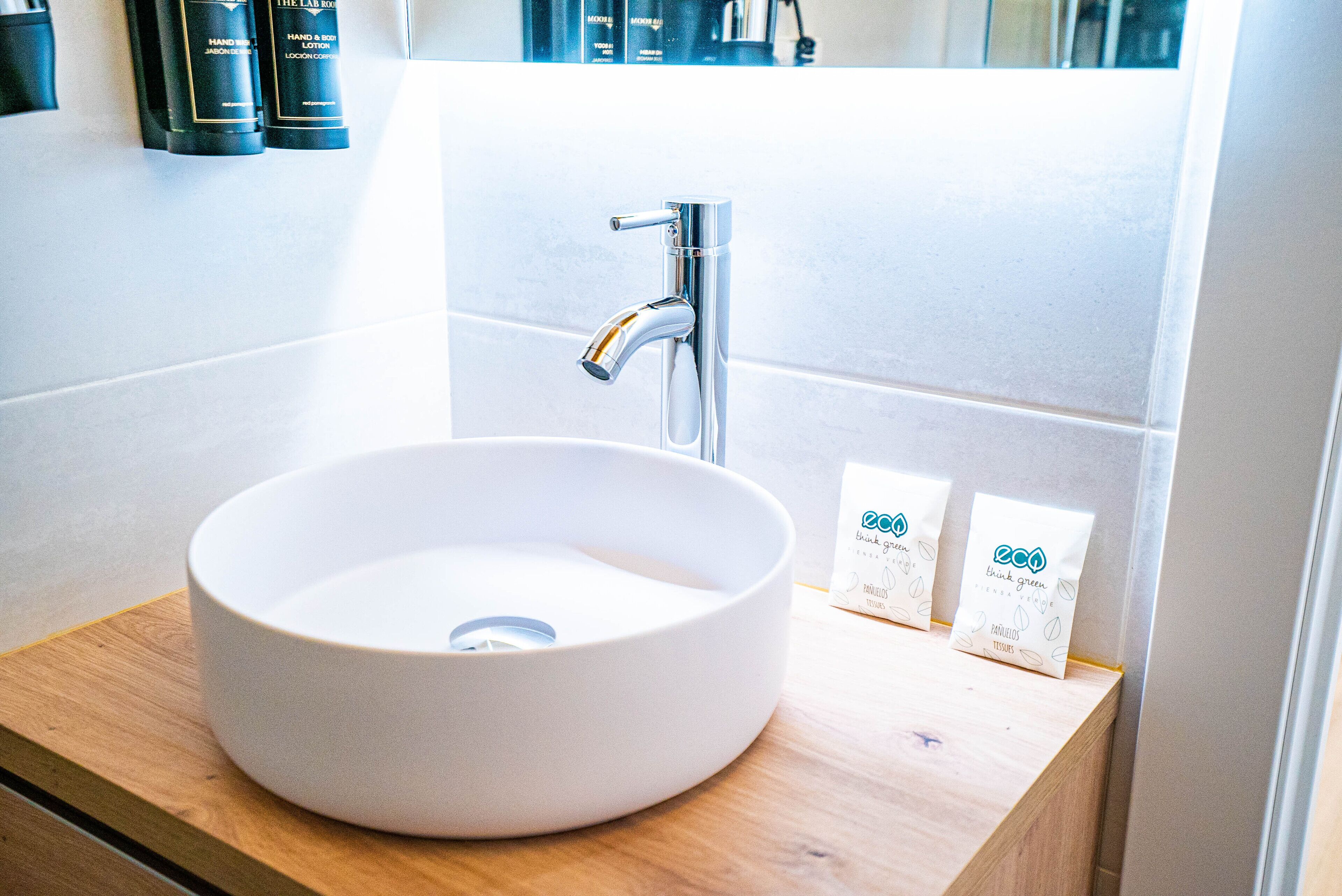 Une salle de bain avec un lavabo blanc, un robinet chromé et un plan de toilette en bois. On y trouve deux petites boîtes blanches sur le plan de toilette, avec du texte vert.