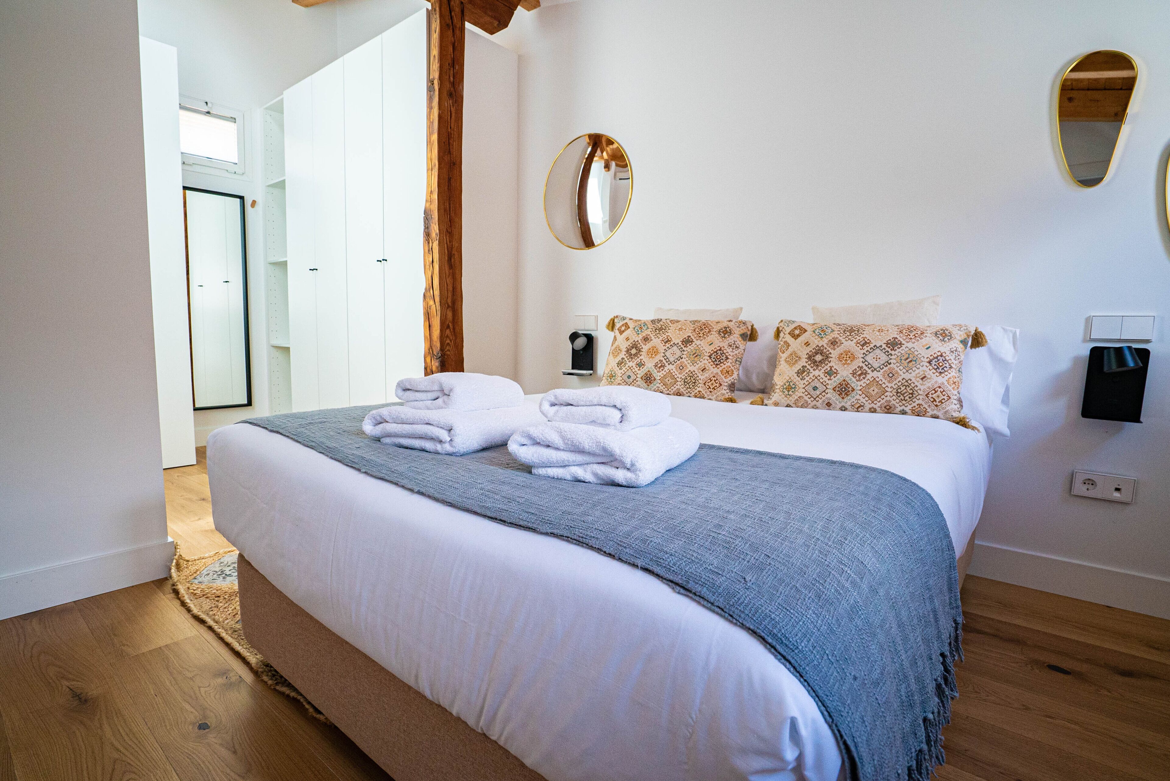 A neatly made bed with white linens and a blue blanket, a wooden floor, and a white door in the background.