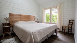 Een slaapkamer met een houten hoofdbord, een groot bed, een nachtkastje met een lamp, een stoel en een raam met gordijnen.