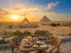 A table with food and drinks, wicker chairs, and a view of the pyramids at sunset. - Tut Pyramids View Hotel (Giza)