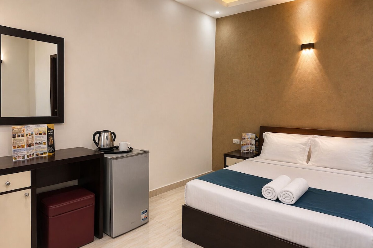 A hotel room with a bed, a desk, a refrigerator, a mirror, and a wall-mounted light fixture.