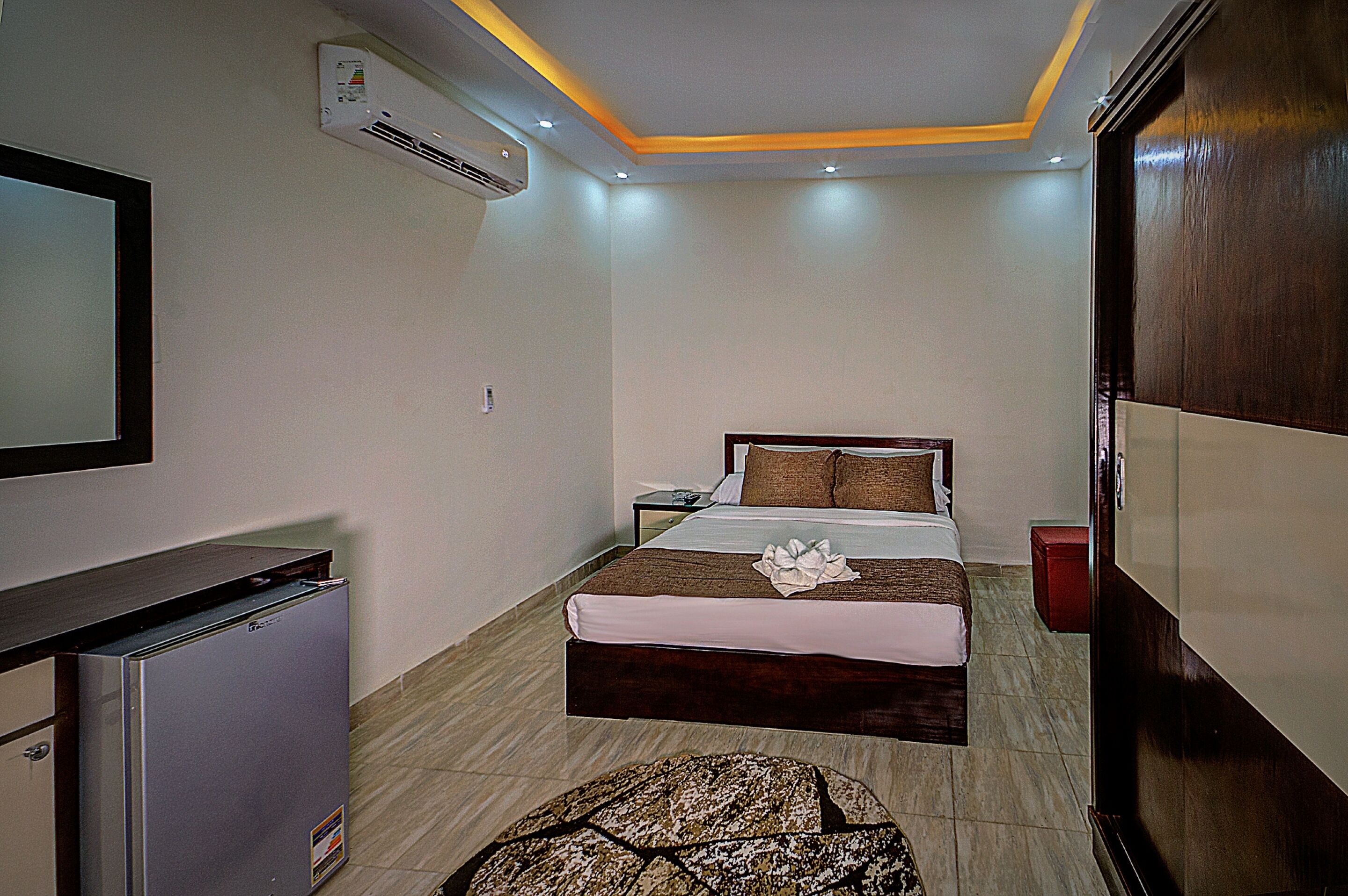 A hotel room with a bed, a small desk, a refrigerator, an air conditioner, and a wardrobe.