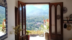 A room with a view of a mountainous landscape through an open door. There are potted plants on the floor and a shelf with books and decorative plates on the wall. - Kasbah de Ouirgane (Ouirgane)