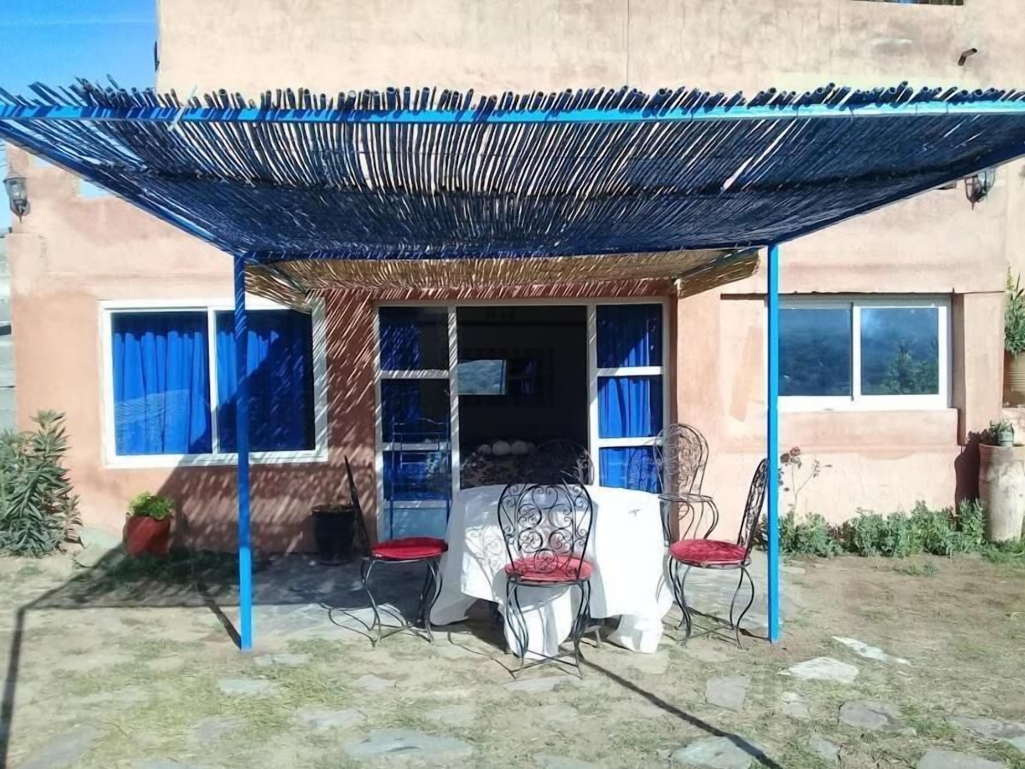 An outdoor patio with a table and chairs, a covered area with a blue canopy, and a building with windows and curtains.
