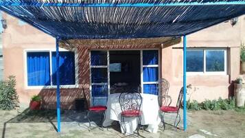 An outdoor patio with a table and chairs, a covered area with a blue canopy, and a building with windows and curtains.