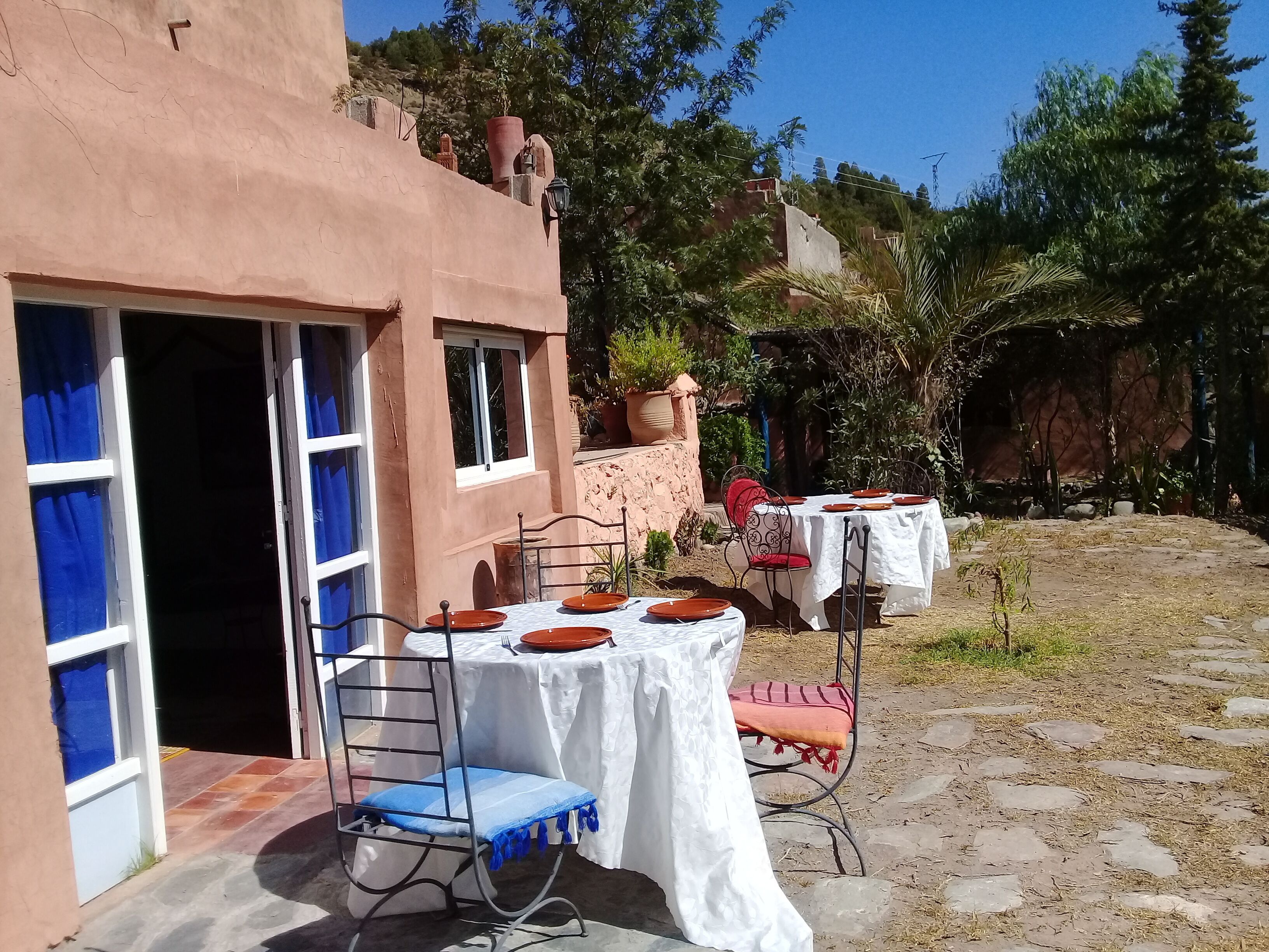 Un espacio exterior con mesas y sillas, la entrada de un edificio y vegetación circundante.