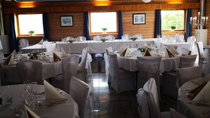 Un salón de banquetes con varias mesas redondas dispuestas para un evento formal, paredes con paneles de madera y grandes ventanas con cortinas.