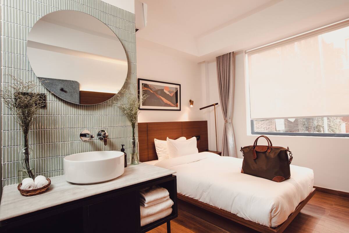 A modern hotel room with a round mirror, a round sink, a bed with white linens, a brown handbag on the bed, and a window with curtains.