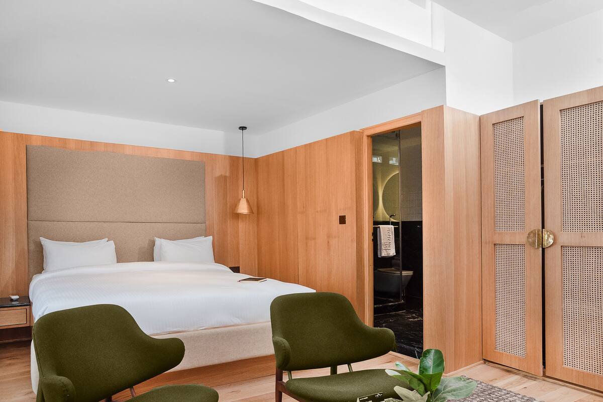 A modern hotel room with a large bed, two green armchairs, a wooden headboard, and a walk-in closet with a mirror.