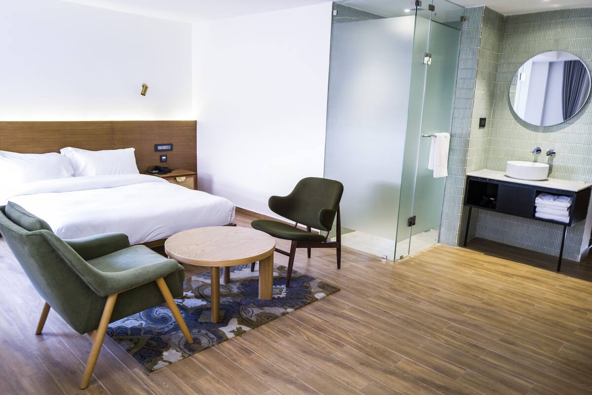 A hotel room with a bed, two chairs, a small table, a bathroom with a sink and mirror, and a shower area.
