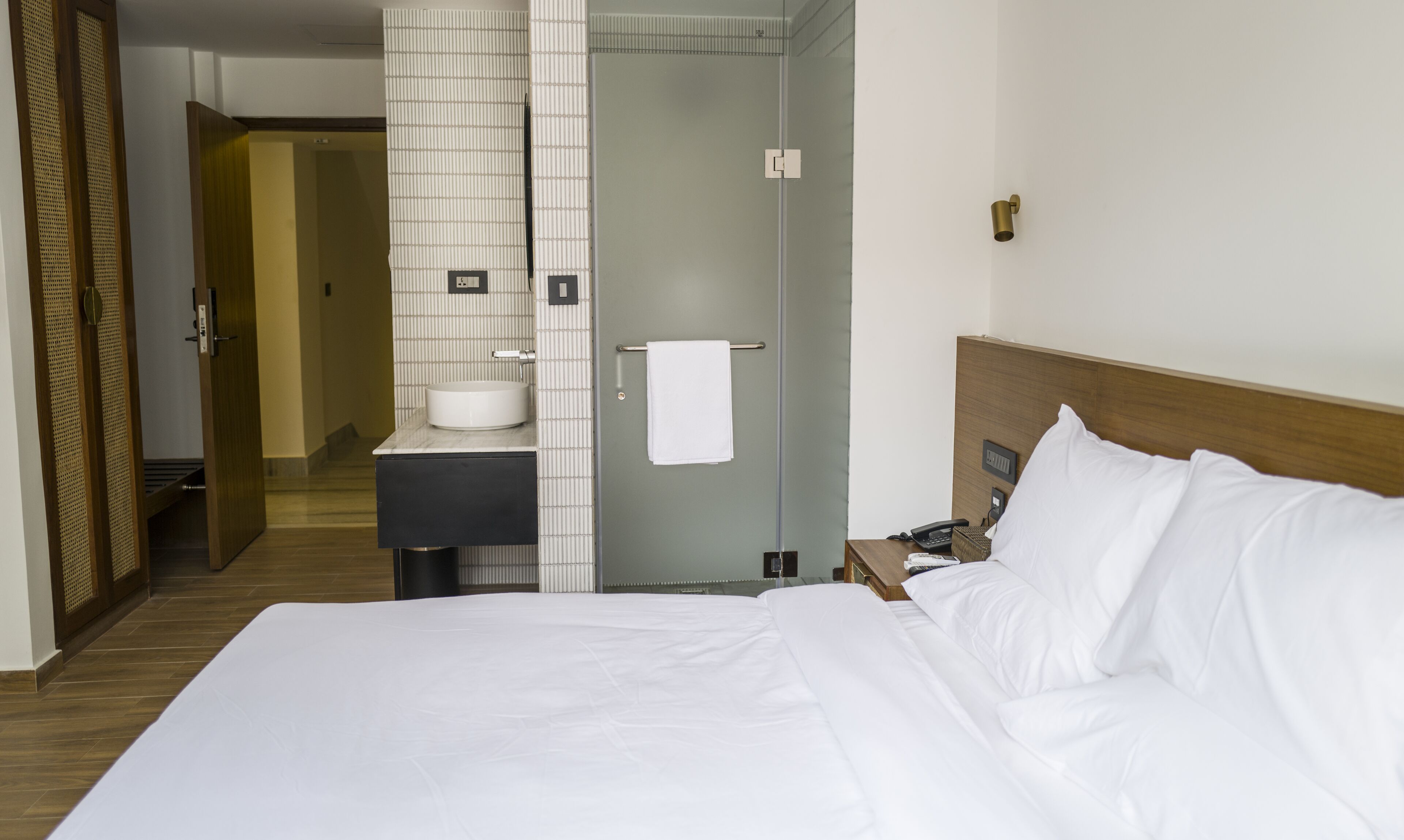 Une chambre d’hôtel comprenant un lit, une table de chevet avec un téléphone, une petite table de chevet avec un panier, une salle de bain avec un lavabo et un porte-serviettes, et un plancher en bois.