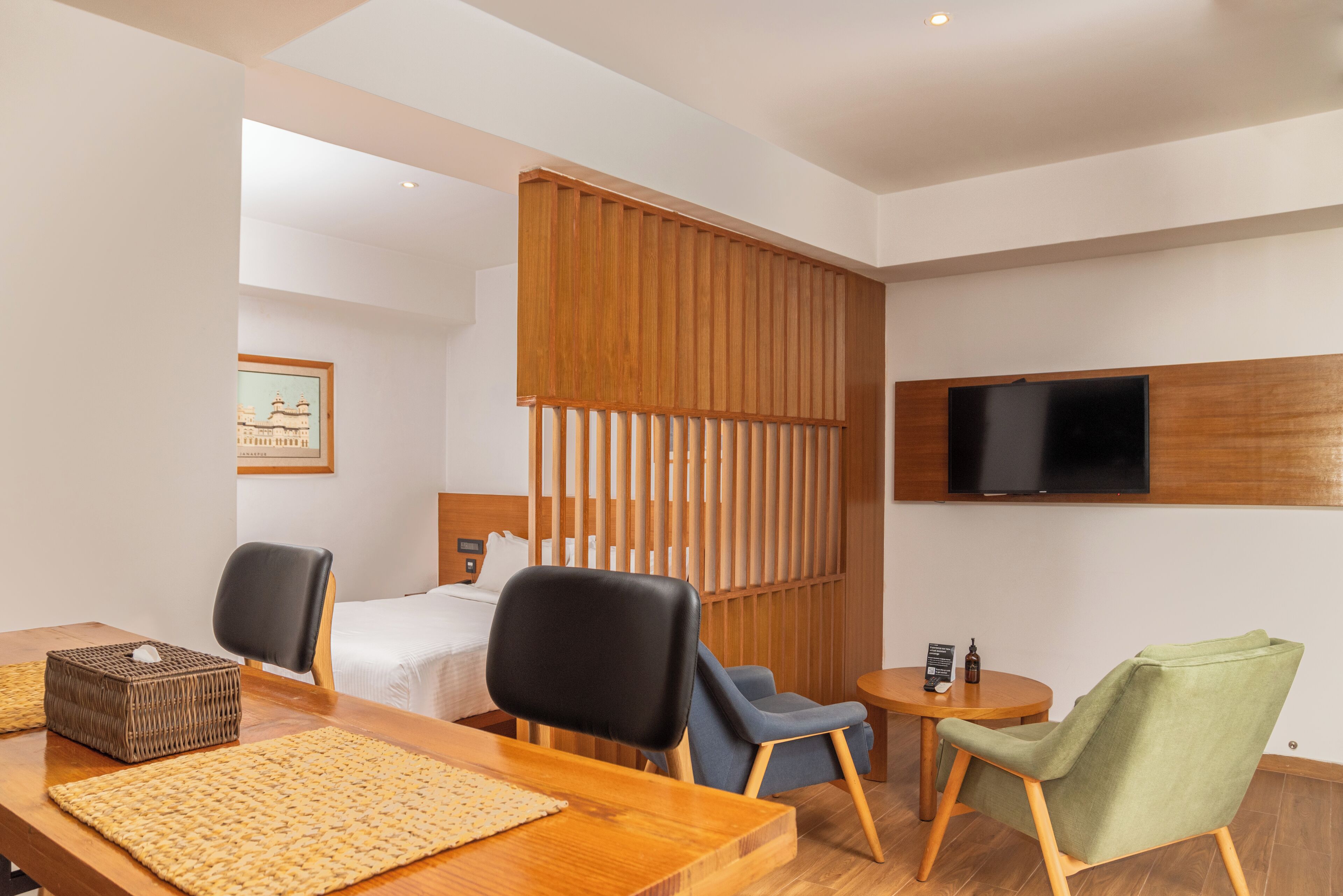 Une chambre d’hôtel avec un lit, un bureau, deux chaises, une petite table avec une bouteille et un téléviseur fixé au mur.