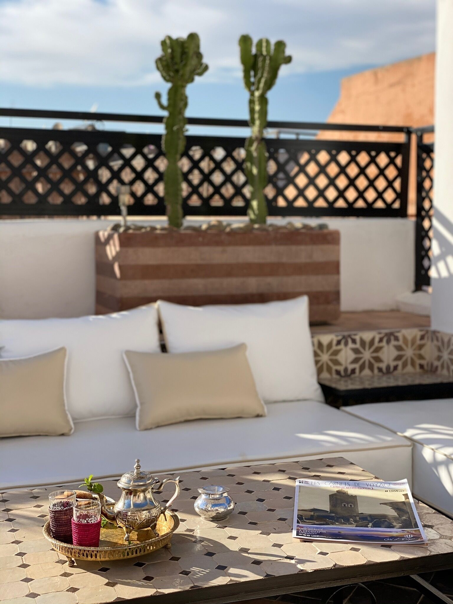A cozy outdoor seating area with a white sofa, cushions, a tray with drinks and a teapot, and two cacti in the background.