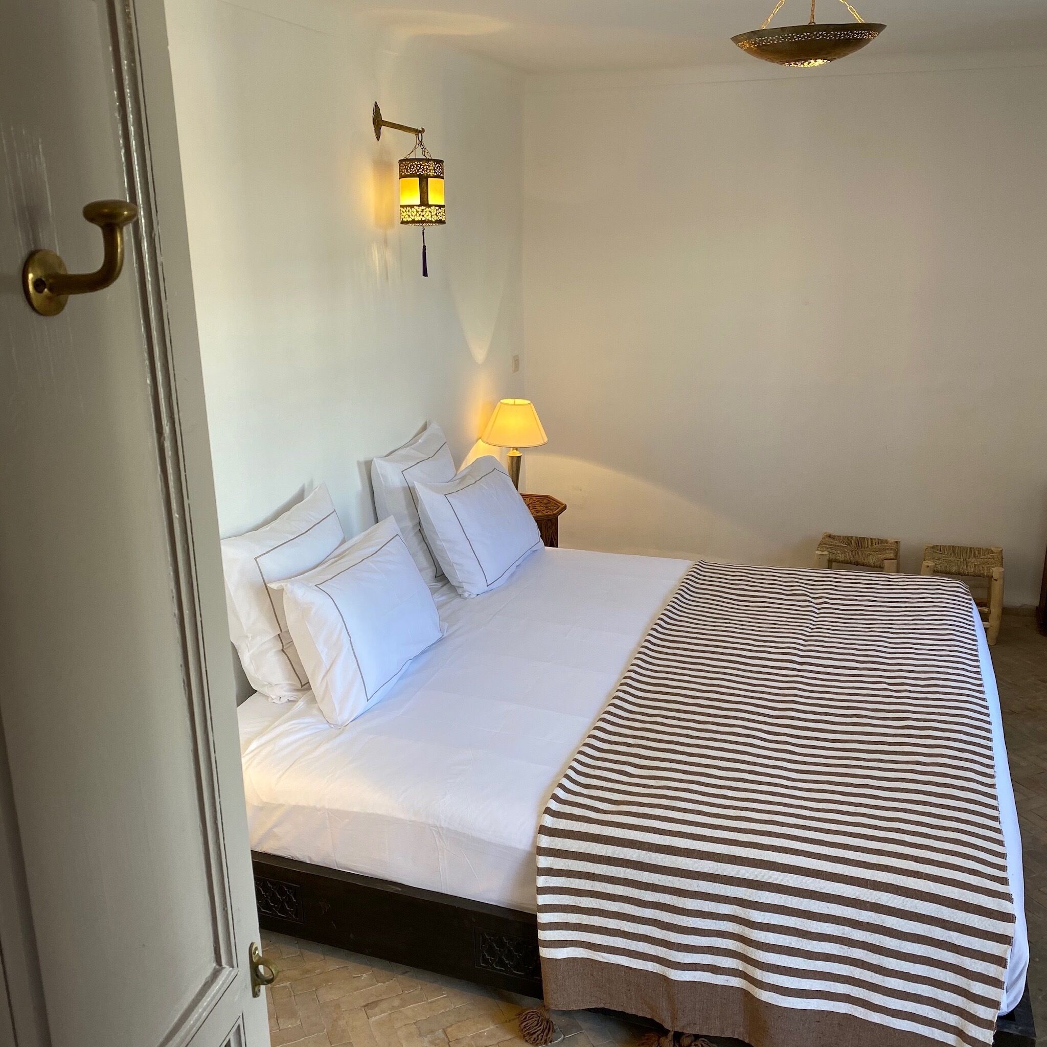 Una cama bien hecha con ropa blanca y una manta a rayas, una lámpara de noche y una lámpara de pared.