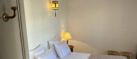 Una cama bien hecha con ropa blanca y una manta a rayas, una lámpara de noche y una lámpara de pared.