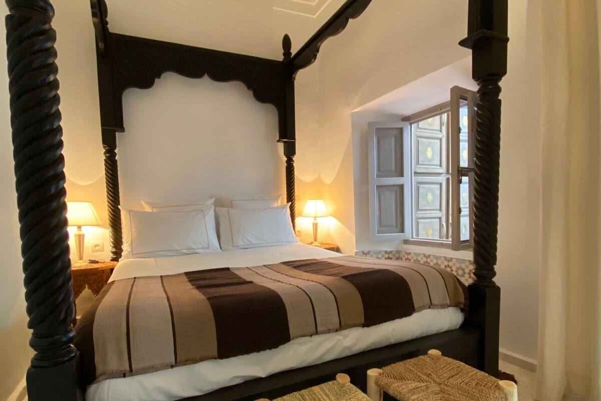 Una cama con dosel con sábanas blancas, dos lámparas de noche y un ventanal con contraventanas.