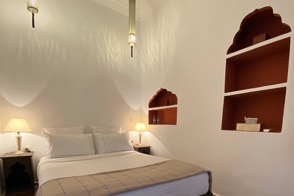 Un dormitorio ordenado con una cama grande, dos mesitas de noche con lámparas, un banco de madera y estanterías empotradas.