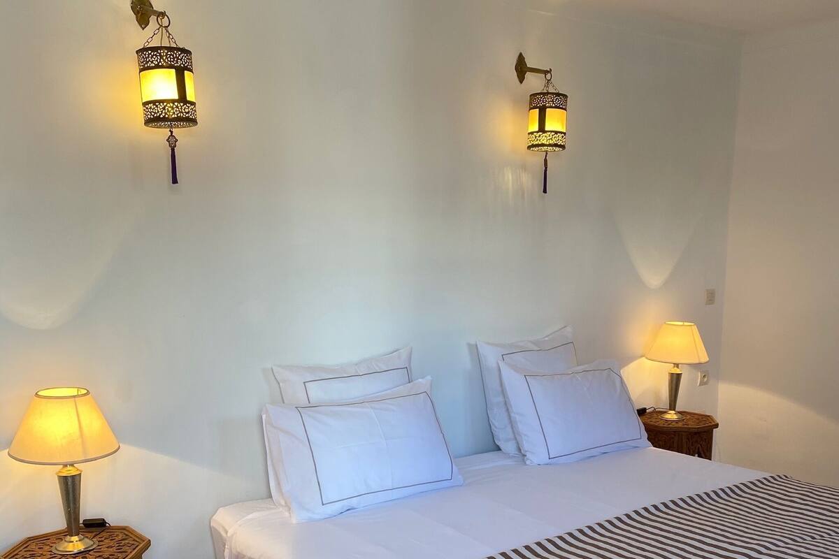 Una cama bien hecha con ropa blanca y cortinas a rayas, dos lámparas fijadas a la pared y una mesita de noche con una lámpara a cada lado de la cama.