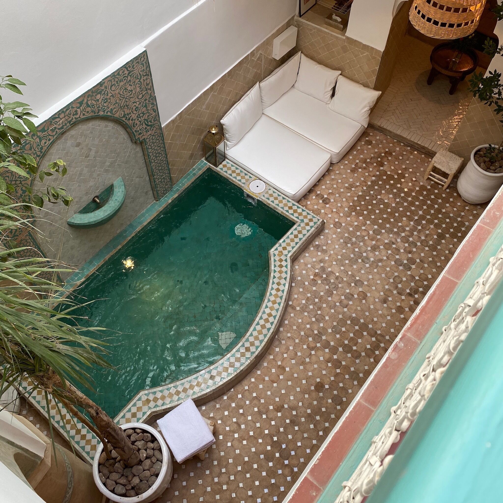 Una piscina pequeña y cercada con borde a cuadros, rodeada por un patio empedrado con un sofá blanco y una pequeña mesa.