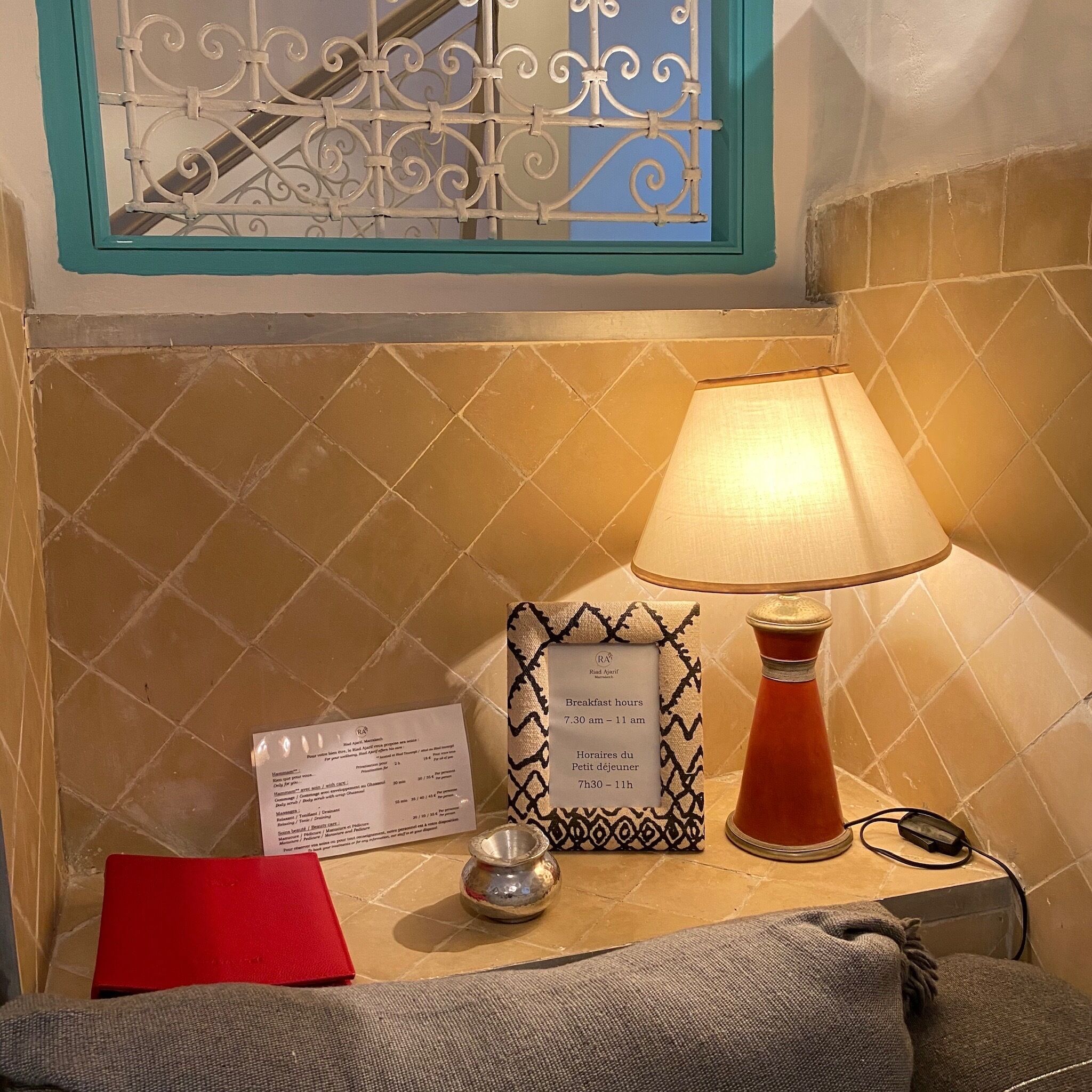 Camera d'albergo con parete rivestita di piastrelle, una finestra con inferriate decorative, un tavolo con una lampada, un menu e una cartella rossa.