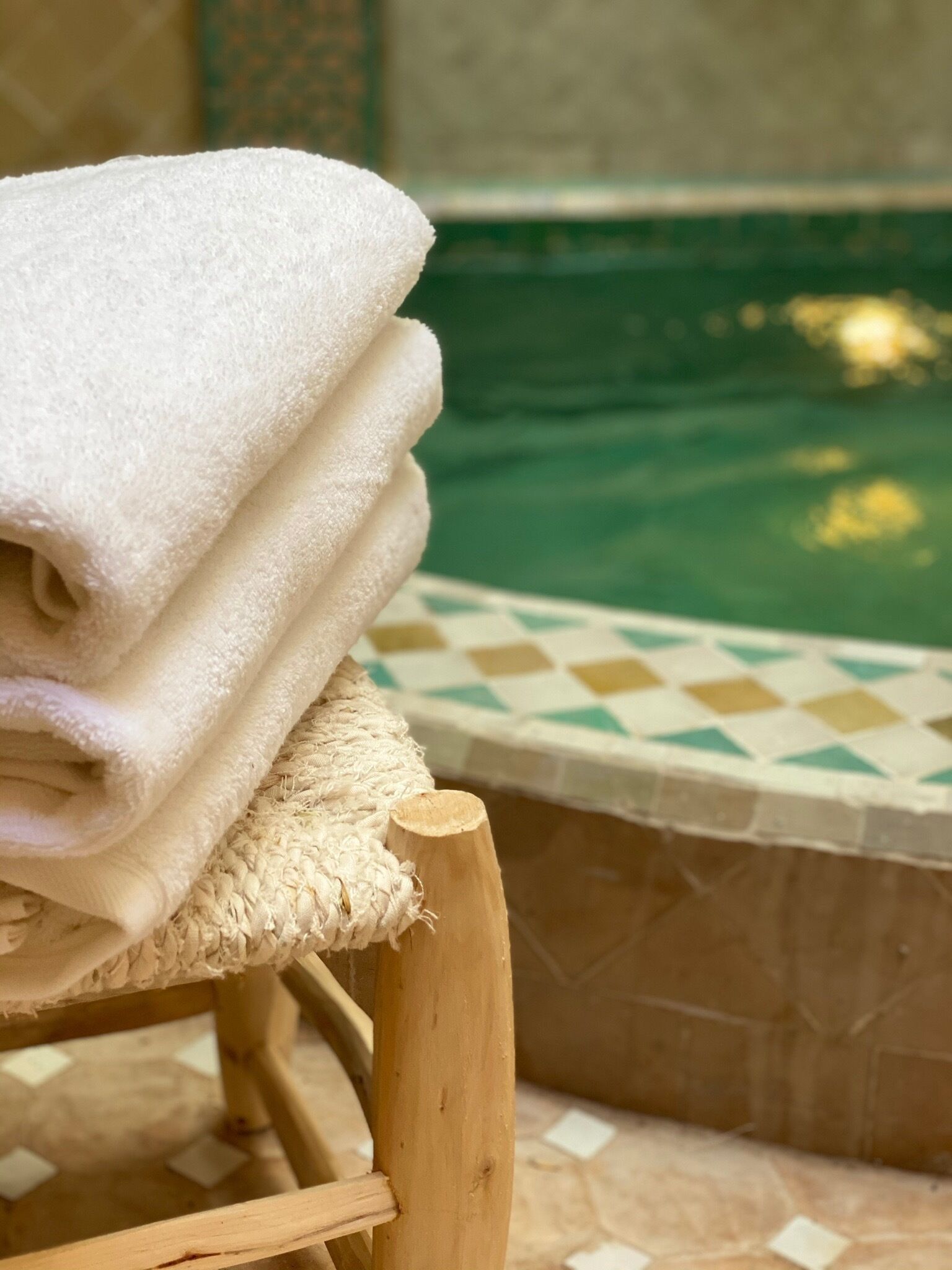 Una pila de toallas blancas sobre una silla de madera junto a una piscina con borde de baldosas a cuadros.