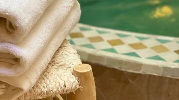 Una pila de toallas blancas sobre una silla de madera junto a una piscina con borde de baldosas a cuadros.