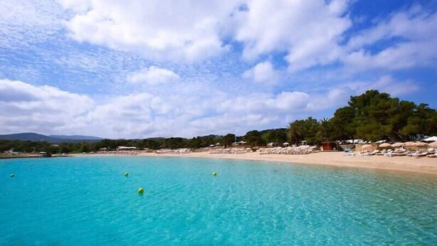 Uma praia com águas turquesa cristalinas, areal arenoso e uma fileira de guarda-sóis e espreguiçadeiras.