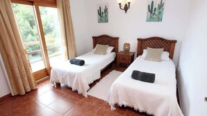 Quarto com duas camas de solteiro, cabeceira de madeira, mesinha de cabeceira com abajur e janela com cortinas.