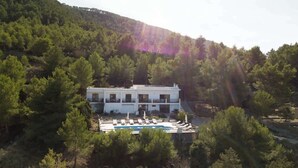 Um edifício branco com piscina e espreguiçadeiras, rodeado por árvores e colinas.