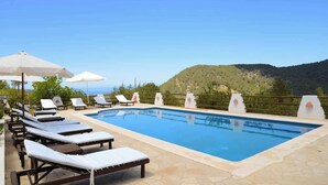 Piscina com espreguiçadeiras e guarda-sóis, rodeada por um deck de pedra e vista para uma paisagem montanhosa.
