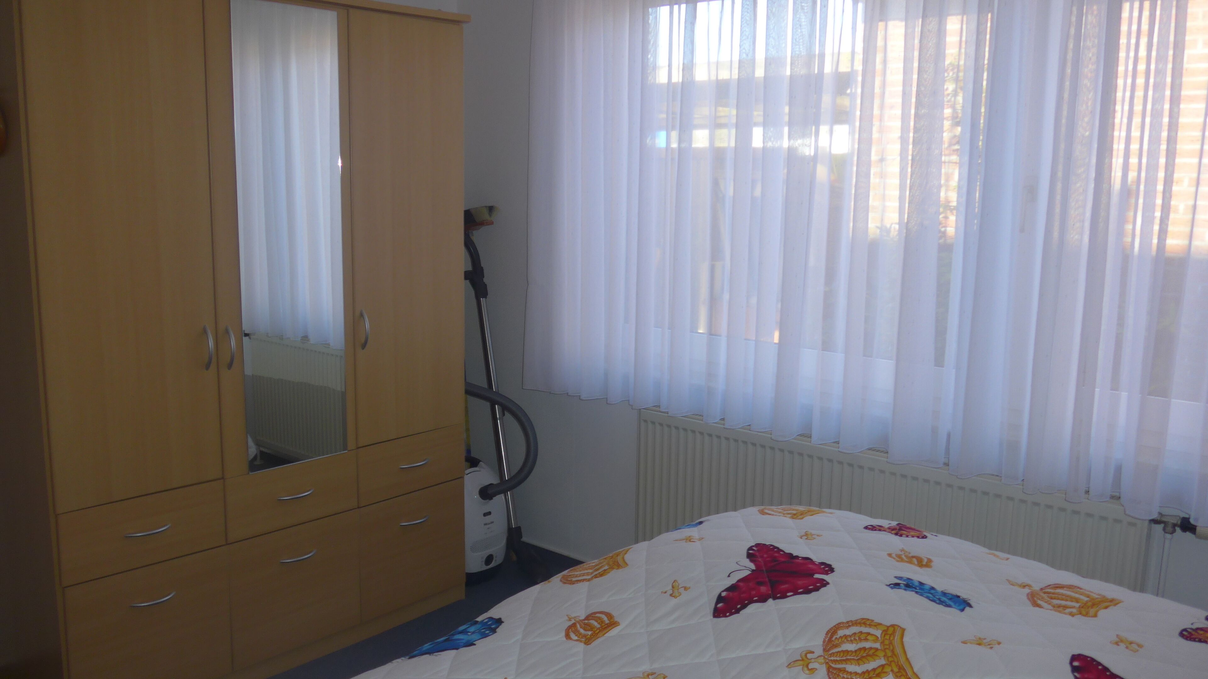 A bedroom with a wooden wardrobe, a bed with a colorful quilt, and a window with sheer curtains.