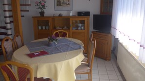 A dining area with a round table covered by a yellow tablecloth, surrounded by wooden chairs with striped cushions. A cabinet with glass doors and a television on top is visible in the background. - Haus Eveline (Borkum)