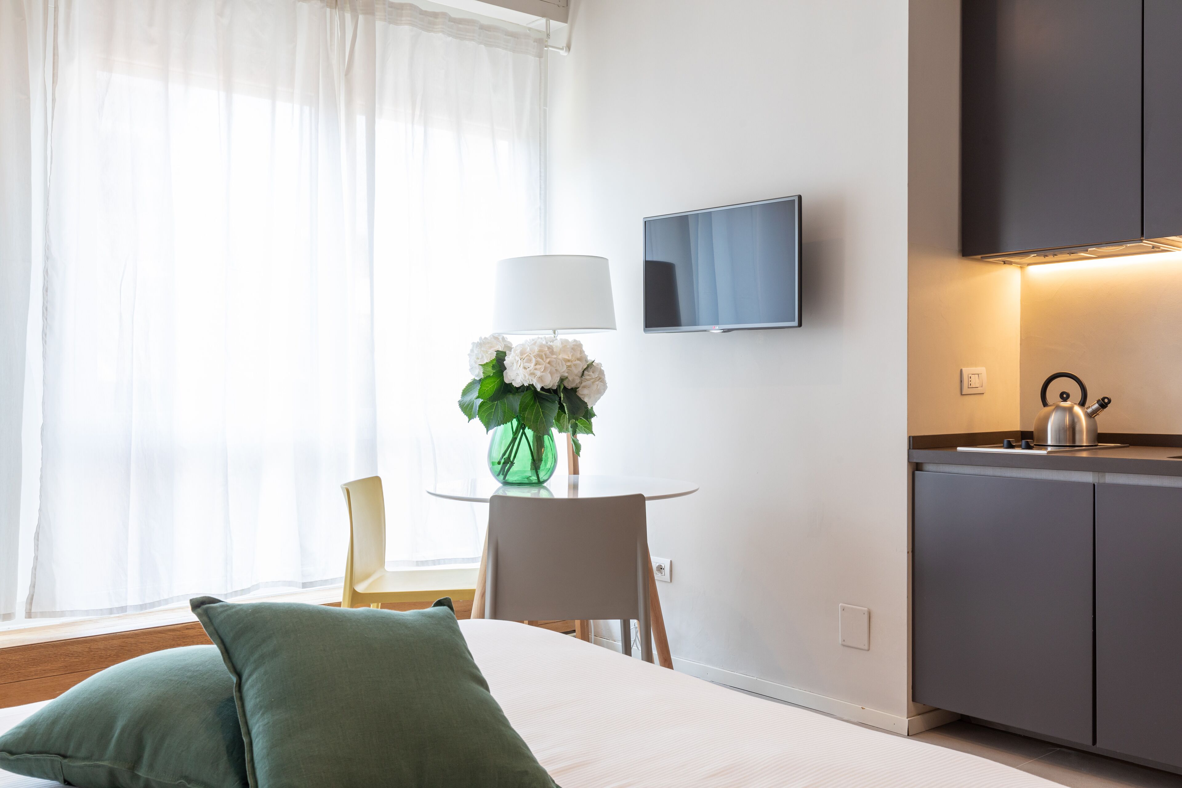 Habitación de hotel con cama, escritorio con silla, televisión montada en la pared y una pequeña cocina.