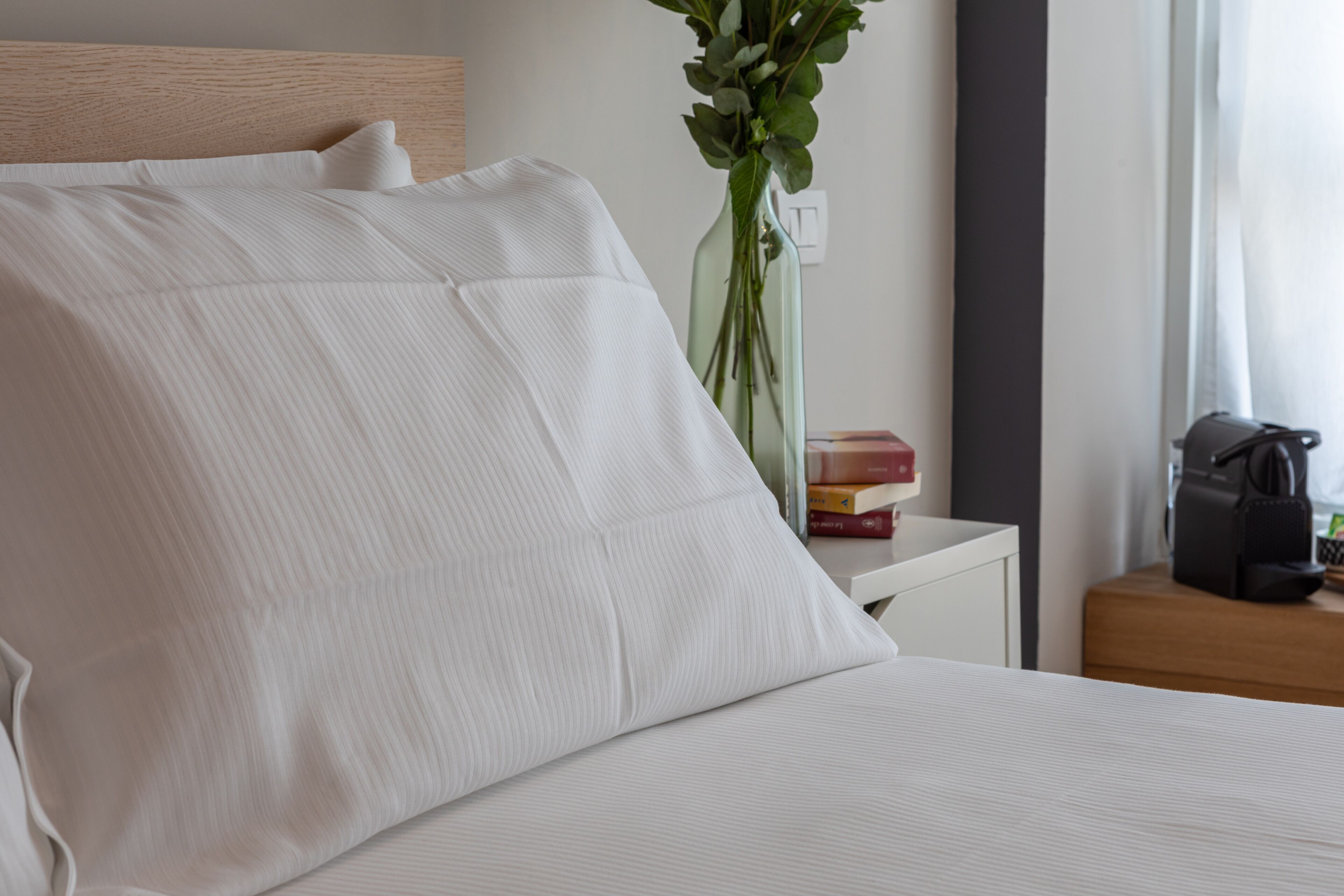 A neatly made bed with white linens, a bedside table with a vase of green leaves, and a few books.
