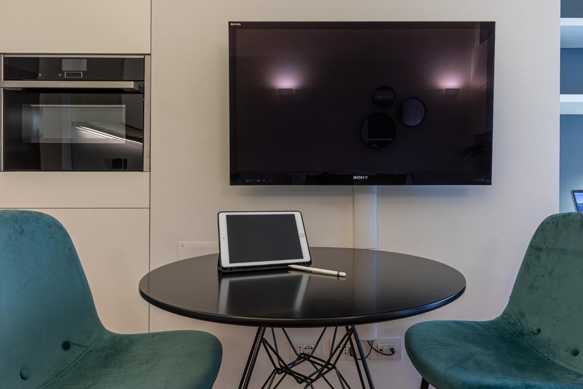 A modern room with a flat-screen TV mounted on the wall, a small round table with a tablet and pen, and two teal chairs.