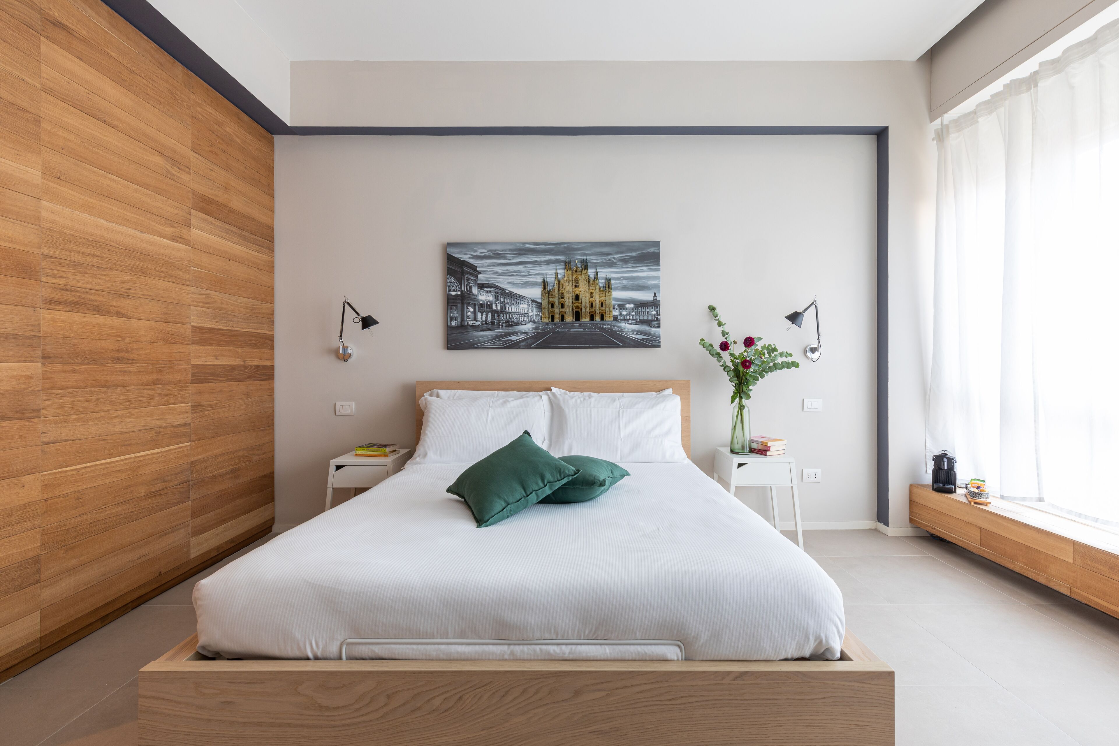 A modern bedroom with a wooden headboard, a large bed with white bedding and green pillows, a cityscape painting on the wall, bedside lamps, and a window with sheer curtains.