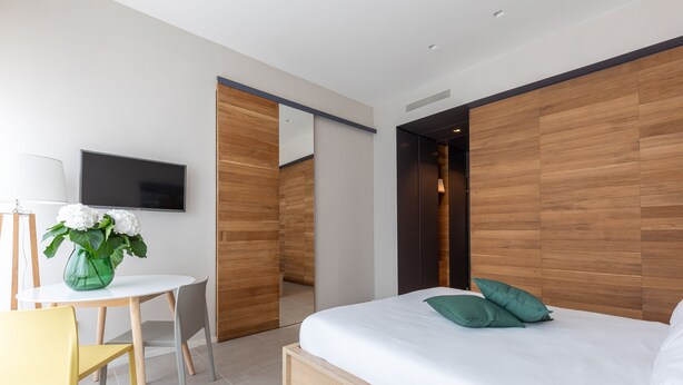 Un dormitorio moderno con cabecera de madera, cama con almohadas verdes, mesita de noche con un florero y un televisor de pantalla plana en la pared.