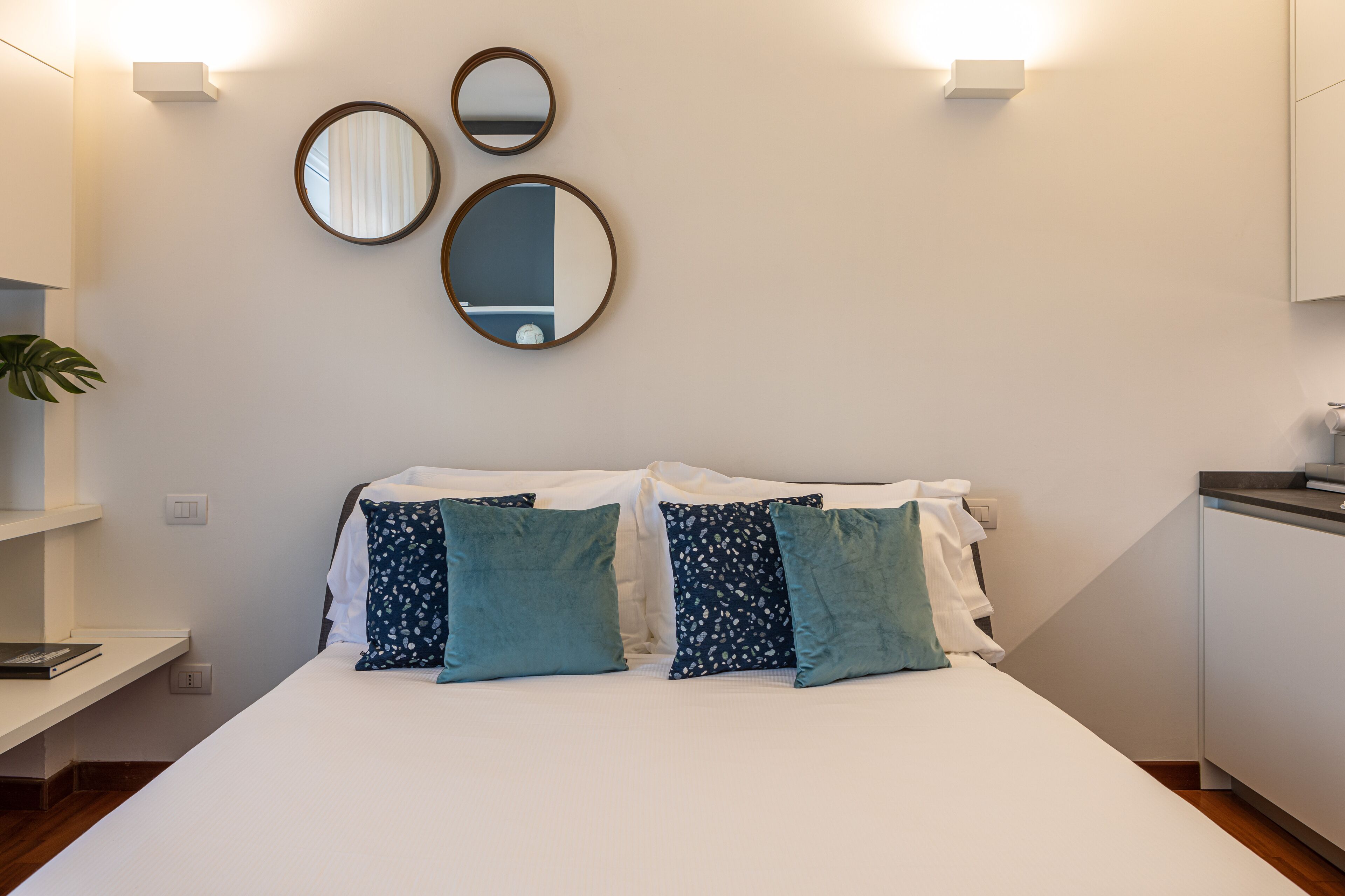 Una cama bien hecha con dos almohadas, una mesita de noche con un libro y un espejo montado en la pared.