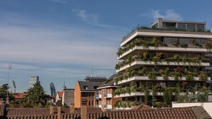 Un edificio de varios pisos con balcones decorados con plantas, cielo despejado y edificios urbanos circundantes.