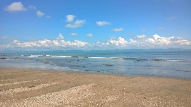 Een strand met kiezelstenen, golven en verafgelegen bergen onder een heldere lucht.