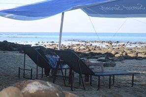 A beachside seating area with two lounge chairs under a canopy, a table with a drink, and a view of the rocky shoreline and ocean.
