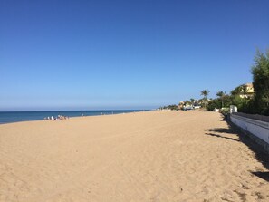 A wide sandy beach with a clear blue sky, palm trees, and buildings in the background. - Attractive bungalow in a quiet residential complex with pool (Dénia)